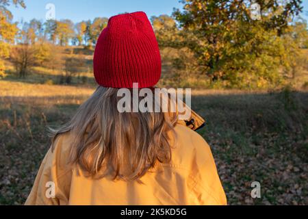 Young woman with long blonde hair in a red hat and yellow coat walking in autumn park. Back view. Bright autumn colors. Sunny weather. Enjoy life Stock Photo