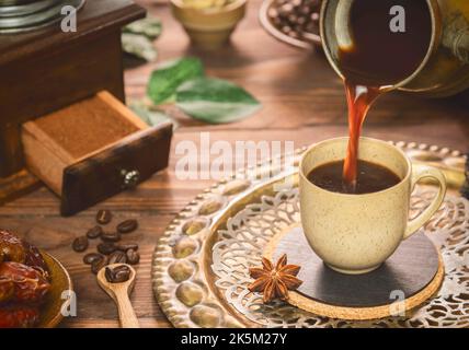 Pouring turkish  coffee into cup.   Cup of coffee placed with roasted coffee beans, fresh dates and ground coffee on rustic wooden background. Stock Photo