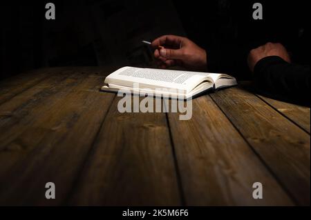 A closeup of a man's hand holding a cigarette and reading a book on a wooden table on dark background Stock Photo