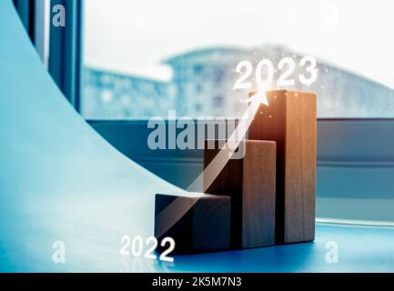 Shining rise up arrow on wood blocks chart steps as a graph from year 2022 to 2023 on blue background with cityscape, business growth process, profit, Stock Photo