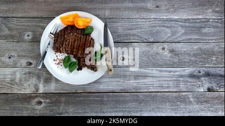 Freshly grilled beef steak with herbs and vegetables on a rustic wood table in flat lay format Stock Photo