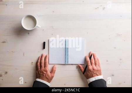 Top view of hands of a businessman holding black marker by an open spiral notebook on wooden desk with a cup of coffee, Stock Photo
