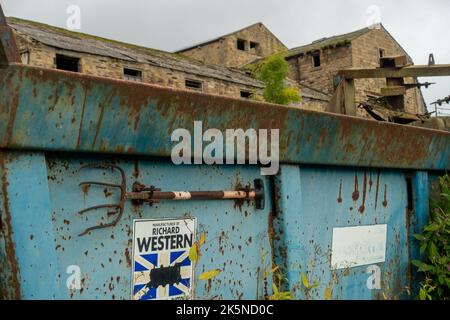 Farmer's pitch fork attached to the side of rusty farm machinery with farm buildings behind, West Yorkshire, England, UK Stock Photo