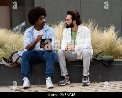 Two diverse young men sitting in a financial district while talking and working on a tablet. Teamwork Stock Photo