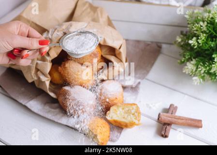Female hand sprinkles powdered sugar on donuts. Cottage cheese round donuts pile covered up with sugar powder, big pile in a paper bag. Whole dough ba Stock Photo