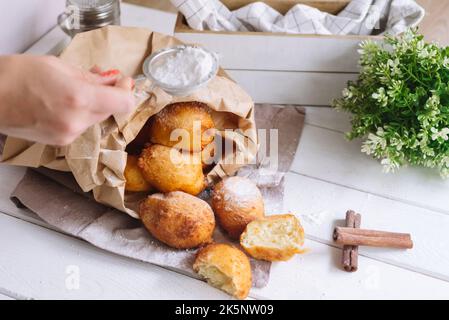 Female hand sprinkles powdered sugar on donuts. Cottage cheese round donuts pile covered up with sugar powder, big pile in a paper bag. Whole dough ba Stock Photo