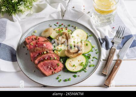 Medium rare meat, almost raw. Sliced roast beef steak with lightly roasted zucchini slices and mushrooms. Homemade dish sprinkled with fresh green oni Stock Photo