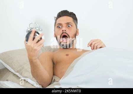 shocked man in bed holding alarm clock Stock Photo