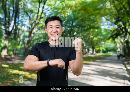 Happy and successful man looking at camera and smiling, asian sportsman satisfied with training result, using fitness bracelet smartwatch, holding hand up celebrating victory, success gesture. Stock Photo