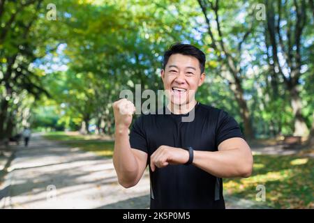 Happy and successful man looking at camera and smiling, asian sportsman satisfied with training result, using fitness bracelet smartwatch, holding hand up celebrating victory, success gesture. Stock Photo