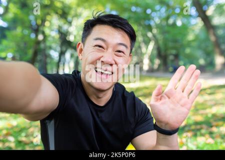 Asian sportsman runner in autumn park looking at smartphone camera and smiling, talking with friends on video call, man in sportswear for fitness in autumn forest. Stock Photo