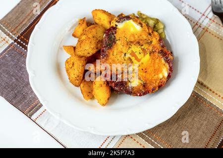 Beef burger with fried egg, baked potato and pickled cucuber on white plate on checkered table-cloth on table, closeup. Stock Photo