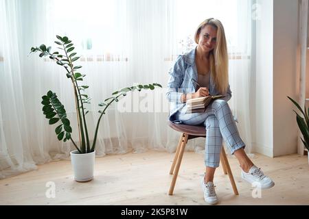 Young woman making notes in notebook sitting on a chair at home,look at camera Stock Photo
