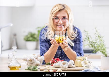 Blonde woman in glasses holds jar of peeled garlic in oil in the kitchen. Stock Photo