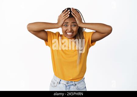 Troubled african american teen girl having big problem, looking frustrated and anxious, holding hands on head and staring nervously at camera, white Stock Photo