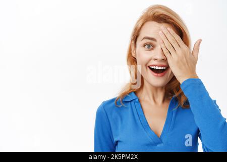 Close up portrait of enthusiastic beautiful woman, 30 years old, she covers one eye and looks amazed, stands over white background Stock Photo