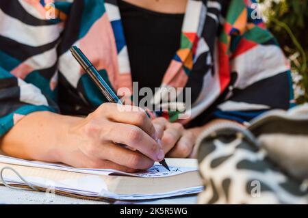 unrecognizable caucasian young woman sitting outdoors drawing with black marker in a notebook, closeup with copy space. Stock Photo