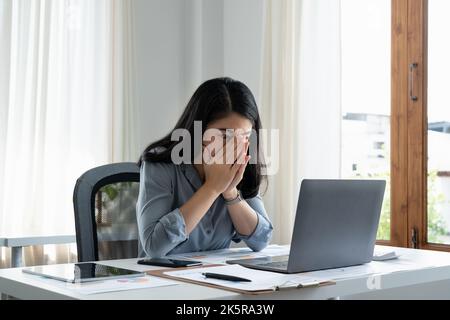 Tired business woman in stress works at a laptop while sitting at a table at home and holds her hand on her temples, migraine attack. Freelance, work Stock Photo