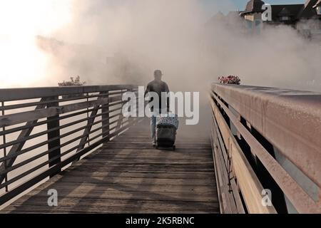 A man walks with luggage across a footbridge as gasses from the natural geothermal hot springs envelope the landscape in Pagosa Springs, Colorado, USA. Stock Photo