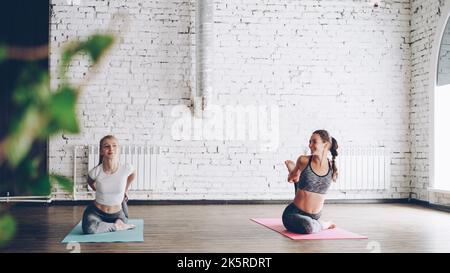 Young blond girl is enjoying individual yoga practice with friendly female instructor in light studio. Women are doing sequence of asanas on bright mats. Friendly relaxing atmosphere. Stock Photo