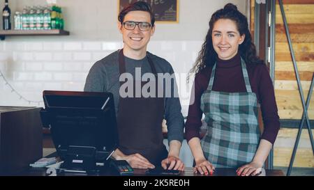 Portrait of two young good-looking waiters standing at cashier's desk in modern coffee-house and smiling. Successful business, happy people and food service concept. Stock Photo