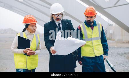 Architect is talking to construction workers showing blueprint walking in building area outdoors. People are wearing safety uniform and helmets. Stock Photo
