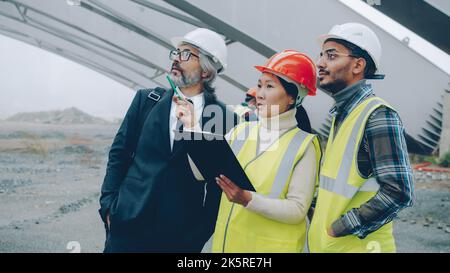 Multiracial group of workers are talking to construction inspector looking at blueprints in building area. Projects and people concept. Stock Photo
