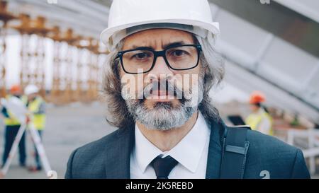 Close-upportrait of serious adult man investor standing at industrial site wearing helmet and looking at camera. Construction and specialists concept. Stock Photo