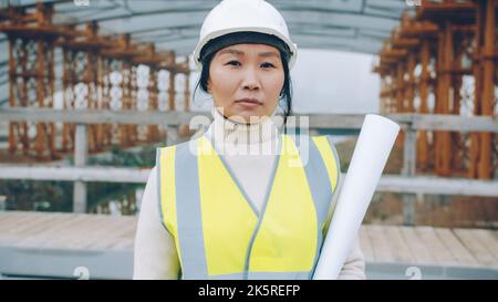 Portrait of successful construction worker Asian woman in helmet and vest holding building plan outdoors in industrial area. People and career concept. Stock Photo