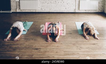 Group of young people are doing yoga exercises moving from King Pigeon pose to Eka Pada Rajakapotasana. Women are concentrated on maintaining position. Studio is light and modern. Stock Photo