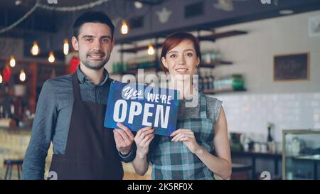 Pretty red-haired businesswoman cafe owner is holding yes we are open sign with her cheerful employee in apron standing near her. Successful start-up and people concept. Stock Photo