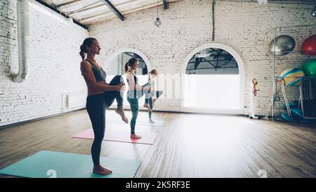 Dolly shot of pretty slim young women doing balancing yoga exercises in light loft style gym. Students are concentrated on practice. Sports equipment, fitness balls and mats are visible. Stock Photo