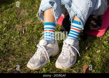 Legs of a girl in stripe bright funny socks and white sneakers. Young woman and black cat sitting on pink mat on green grass. Top view Stock Photo