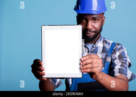 Serious construction worker holding electronic device with blank screen for advertising in front of camera. Builder with digital tablet copy space in studio shot against blue background. Stock Photo
