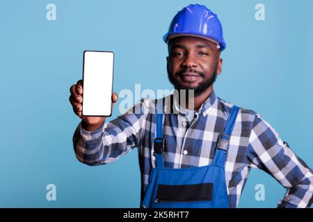 Construction worker in blue uniform work coveralls holds cell phone with empty screen for advertising isolated on blue background. Male professional builder in hard hat looking at camera. Stock Photo