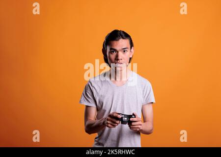 Young asian man playing computer video games with joystick, entertainment activity, fun pastime. Concentrated teenager holding console wireless controller, enjoying videogames Stock Photo