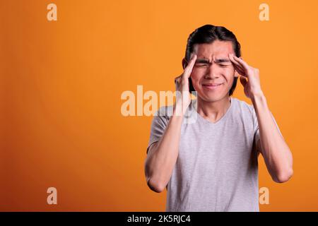 Young man having headache, touching temples with fingers. Exhausted asian teenager with closed eyes suffering from painful migraine symptoms, overworked stressed person feeling unwell Stock Photo