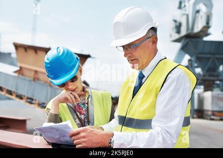 Two heads are always better than one. Two dock workers holding paperwork while standing in the shipyard. Stock Photo