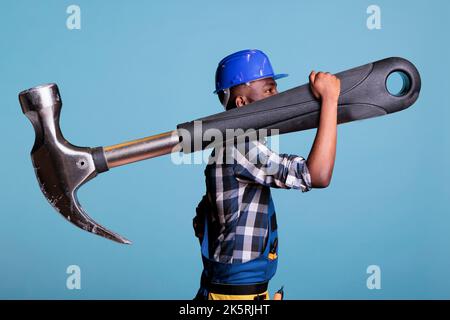 Concept of heavy work, African american builder with giant hammer in studio shot. Strong man heavy work Construction worker carrying huge work tool wearing coveralls and hard hat, construction industry concept. Stock Photo