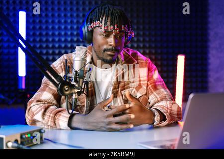 African radio host sitting at desk recording in studio with microphone and laptop Stock Photo