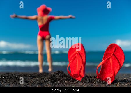 Red flip flops in the black sand and silhouette of woman in red swimsuit standing on the ocean beach. Summer concept. Stock Photo