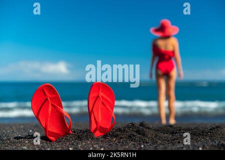 Red flip flops in the black sand and silhouette of woman in red swimsuit standing on the ocean beach. Summer concept. Stock Photo