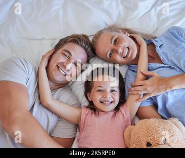 Adorable little girl pulling her parents close while lying in between her mother and father, from above. Faces of loving parents bonding with their Stock Photo