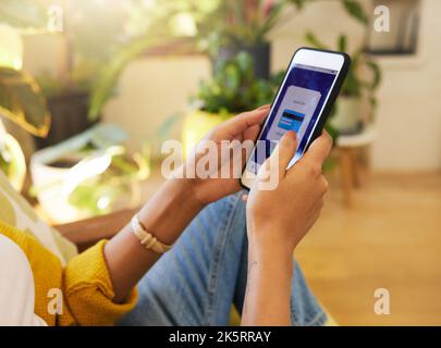 Closeup of a woman banking online while holding her smartphone sitting in a bright living room. A hispanic young female at home using modern Stock Photo