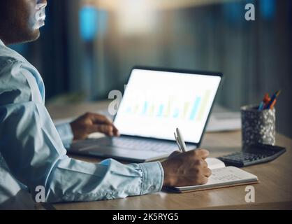 Closeup young african man working on a laptop. African american business man writing in his diary while using a computer late at night in his office Stock Photo