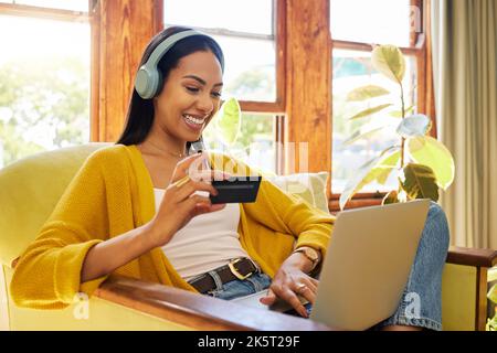 Hispanic woman shopping online while typing on her laptop and holding her bank card sitting on a chair in a bright living room in front of a window. A Stock Photo