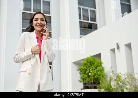 Successful and cheerful young Asian businesswoman having a good conversation on the phone, talking on the phone with someone while walking along the b Stock Photo