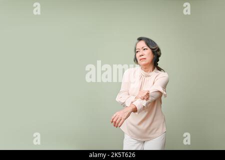 A distressed elderly Asian woman suffering from arm pain, massaging her arm, holding her elbow, standing against the green studio background. copy spa Stock Photo
