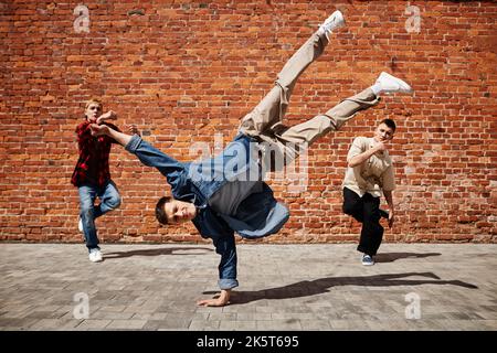 Freeze frame of male breakdance performer doing handstand pose with team against brick wall outdoors Stock Photo