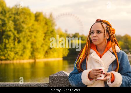 Young woman with dreadlocks in warm clothes standing near lake drinking coffee in park. Stock Photo